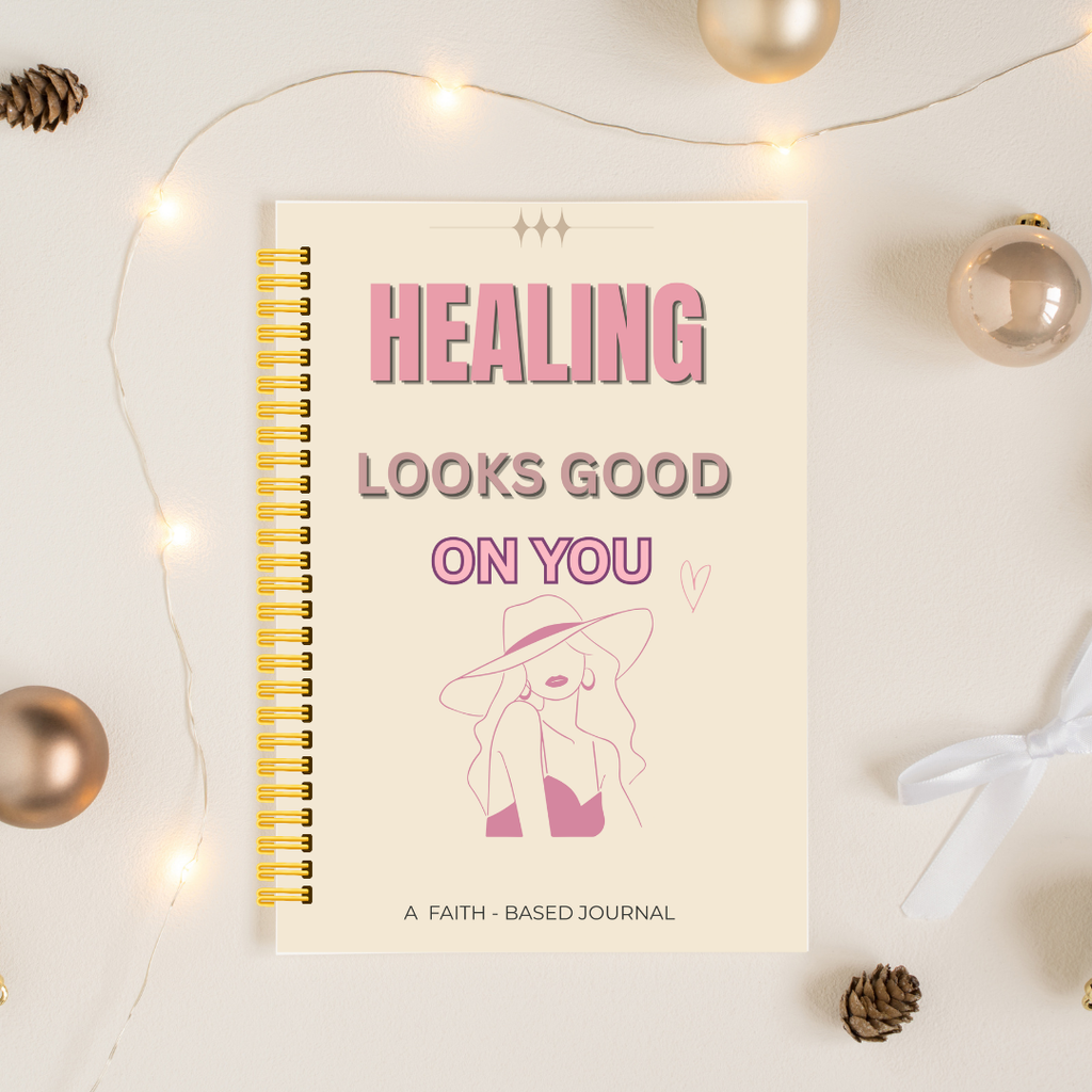 A spiral-bound journal with a cream cover and pink text reading 'HEALING LOOKS GOOD ON YOU' is photographed on a light surface with warm fairy lights, metallic ornaments, pinecones, and a white ribbon around it.