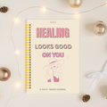 A spiral-bound journal with a cream cover and pink text reading 'HEALING LOOKS GOOD ON YOU' is photographed on a light surface with warm fairy lights, metallic ornaments, pinecones, and a white ribbon around it.