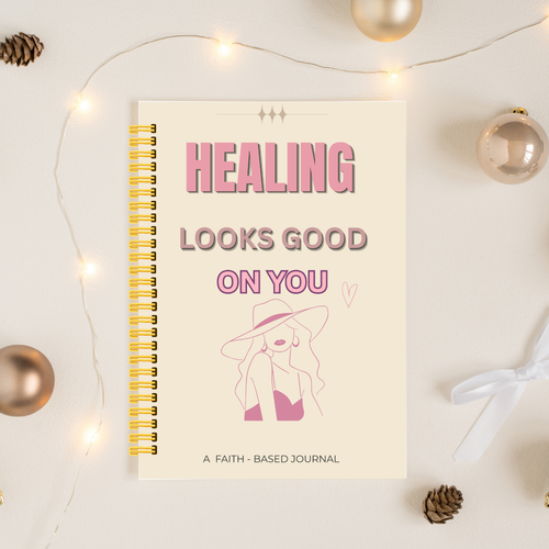 A spiral-bound journal with a cream cover and pink text reading 'HEALING LOOKS GOOD ON YOU' is photographed on a light surface with warm fairy lights, metallic ornaments, pinecones, and a white ribbon around it.